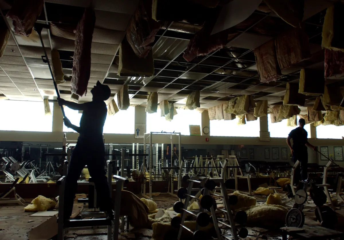 Workers pulling water-soaked ceiling tiles and insulation from a gym ceiling grid after a hurricane