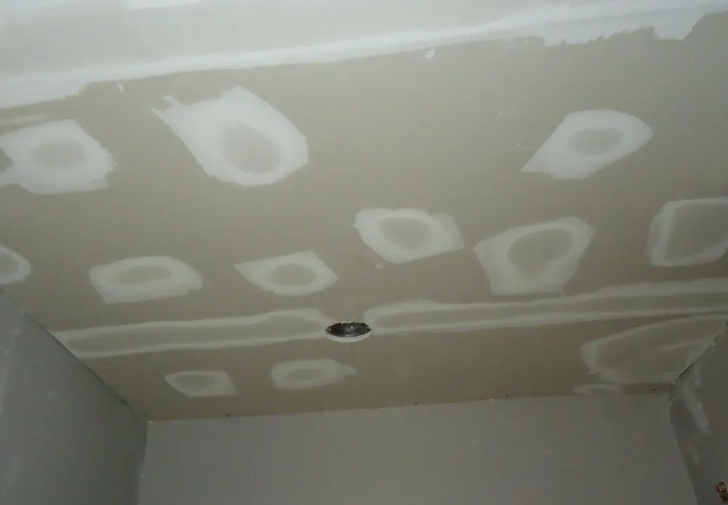 A drywall ceiling during renovation with white joint compound patches over fastener holes and strips of compound along the taped seams between boards