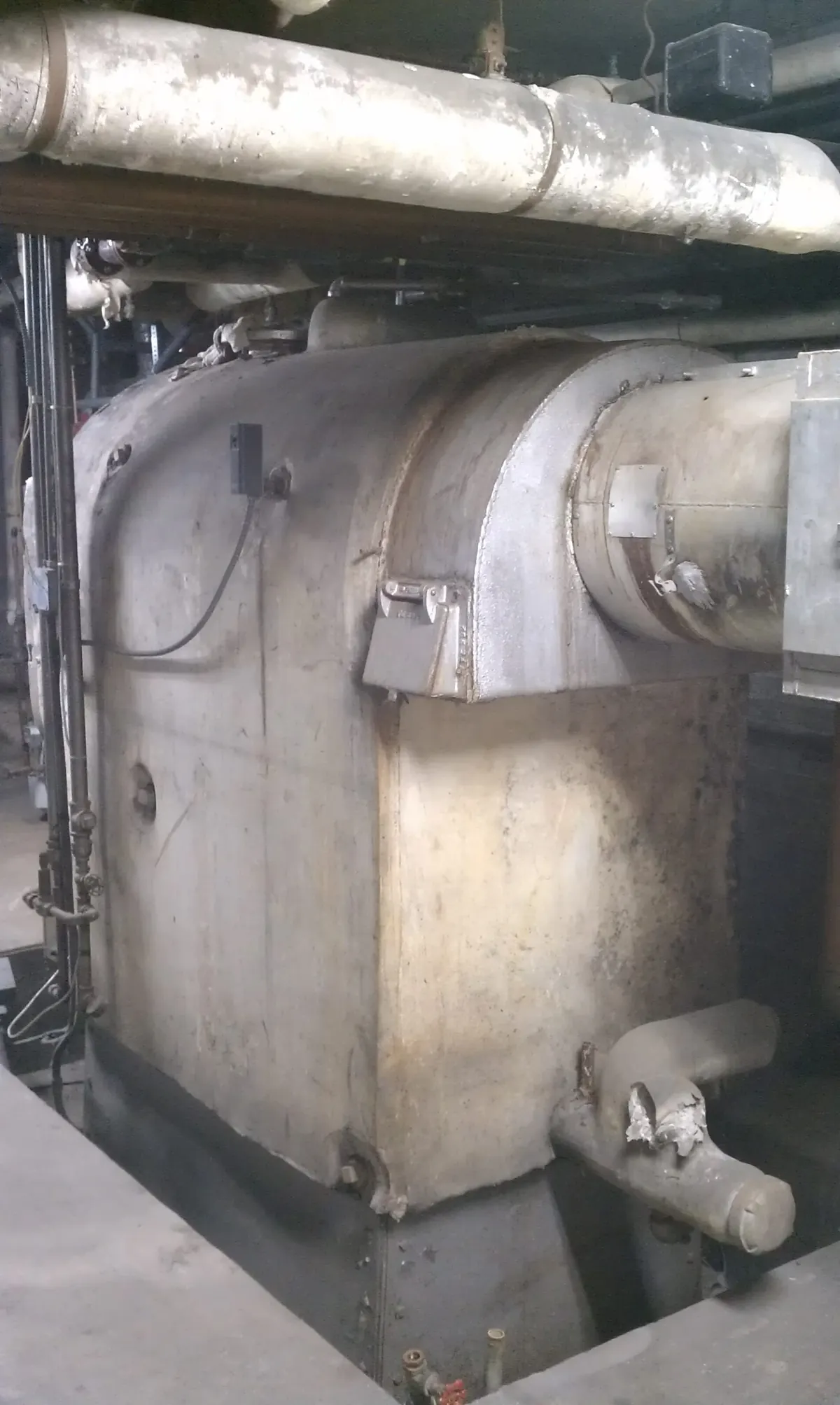 An old basement boiler with a coated shell and jacketed overhead pipe runs, with torn wrap at a pipe end