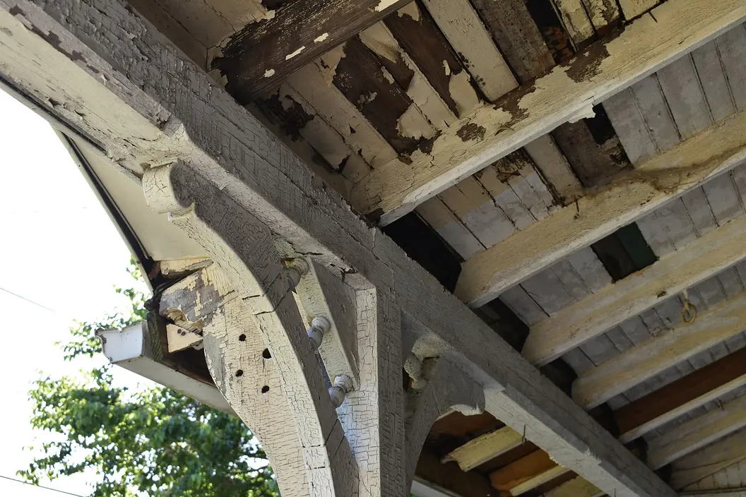 The underside of an old porch roof, with white and grey paint alligatored, cracked and flaking off brackets, beams and ceiling boards