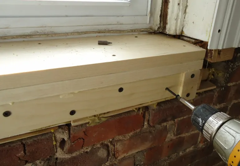 A new pine window sill being screwed into place under an old painted window frame above a brick wall