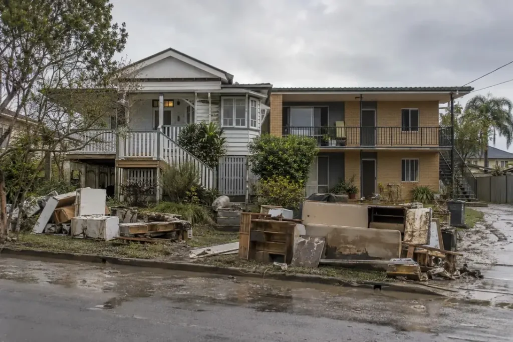 Illustration: an owner-occupied house and a small rental block side by side, both with flood debris at the kerb.