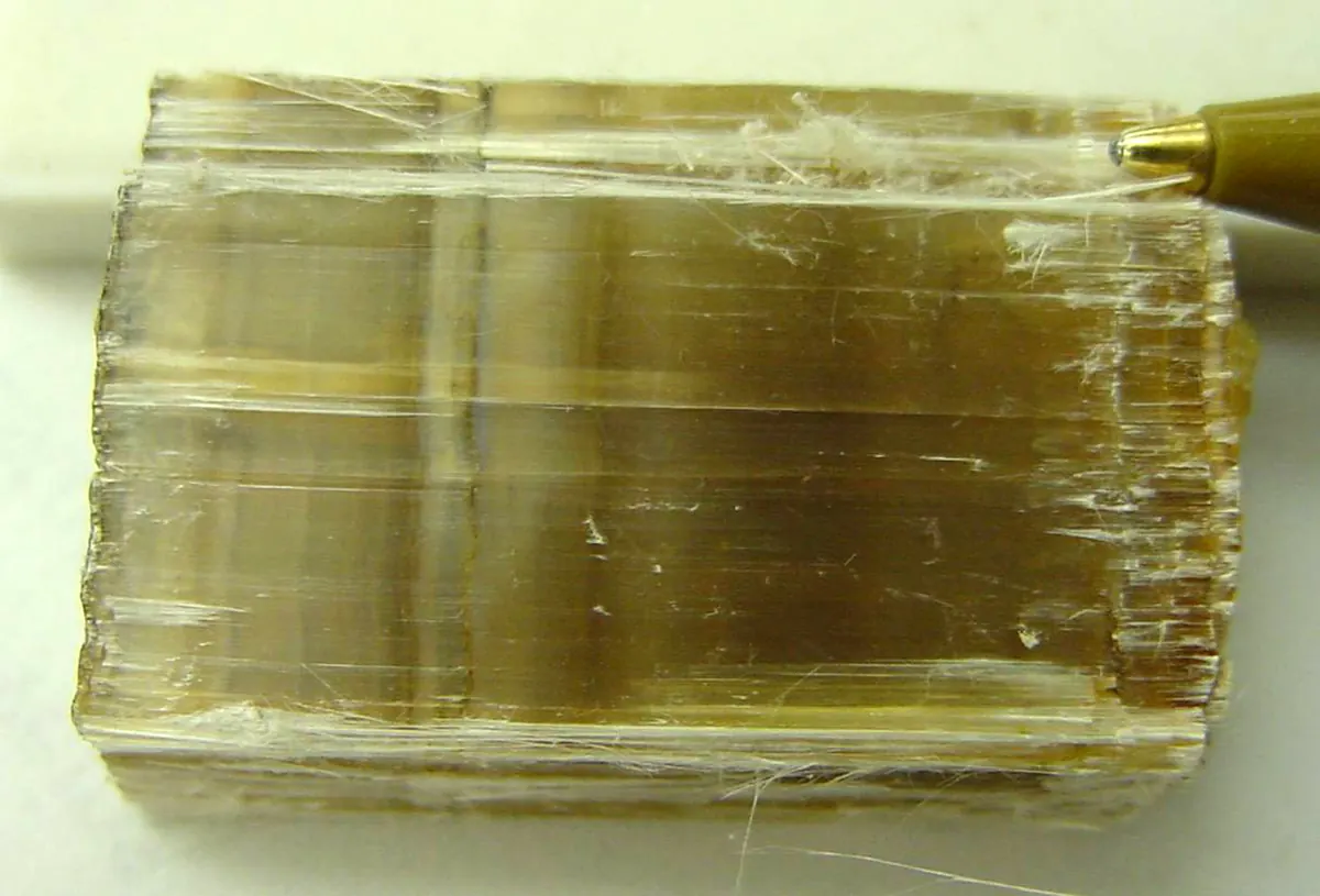 A block of chrysotile with fine silky fibres running across it and loose white fibres lifting at the edge, a pen tip for scale